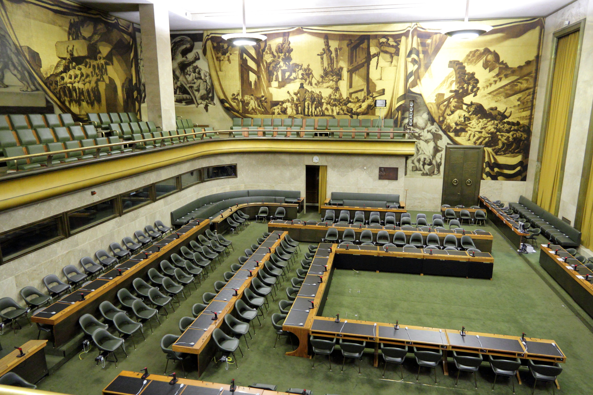 Large empty conference hall with green carpet and U-shaped seating arrangement, featuring rows of microphones and chairs. A gallery with additional seating runs along the upper level. Dramatic mural panels depicting historical and allegorical scenes cover the walls.